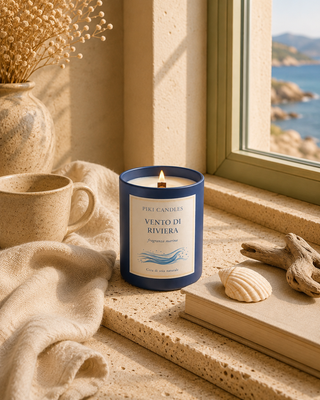 Candela Vento di Riviera Piki Candles accesa su davanzale con vista mare, conchiglie e legni